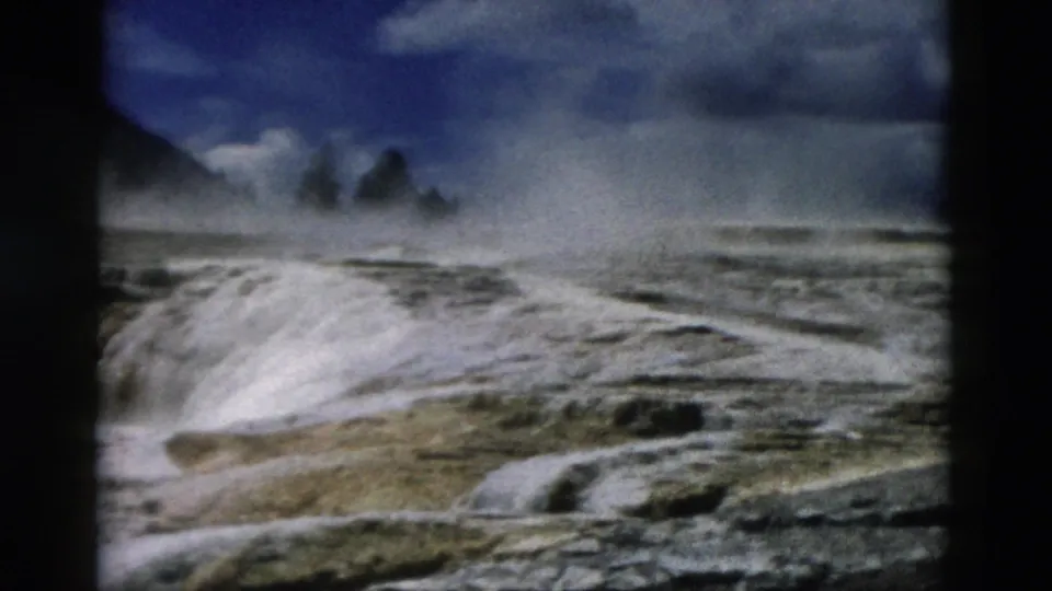 1958 8mm Yellowstone frame showing a pale geothermal mound, rising steam, and dark hills beyond.