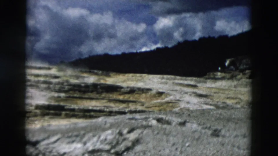 1958 Yellowstone 8mm frame looking across pale terraces and rough rock toward a dark ridge beneath a clouded sky.