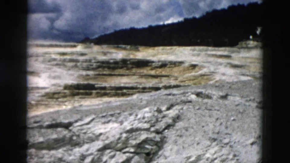 1958 Yellowstone 8mm frame showing rough rocks in front of a pale geothermal basin, distant steam, and a dark tree-covered slope.