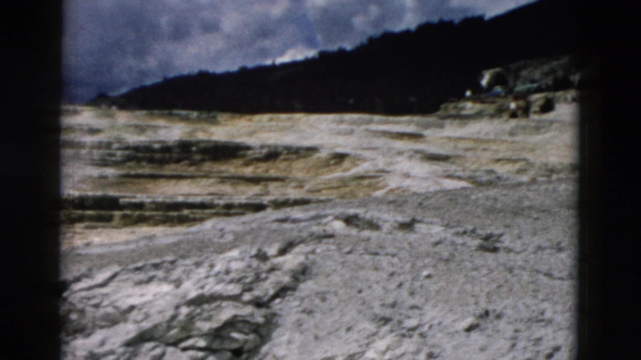 Pale rocky Yellowstone terrain with dark wooded hills under a partly cloudy sky.