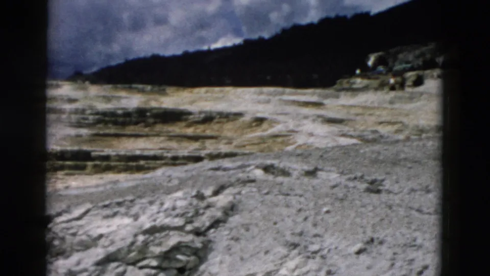 1958 8mm Yellowstone view of pale rocky ground, distant forested hills, and clouds, framed by dark film edges.