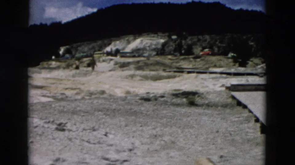 1958 8mm Yellowstone frame showing pale ground, distant wooded slope, and small indistinct roadside or landscape details.