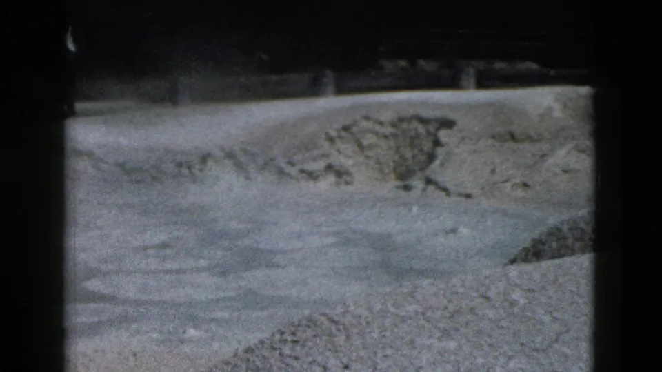 1958 Yellowstone home-movie frame showing pale geothermal ground, rocky deposits, dark frame edges, and tiny distant visitors near the upper edge.