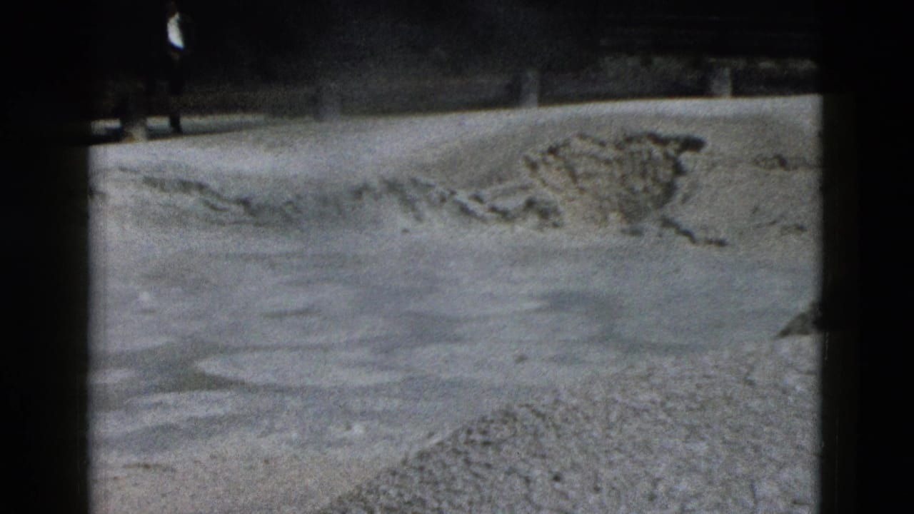 1958 Yellowstone frame with pale mineral terrain, scattered rock deposits, dark border shadows, and a small distant visitor near the top.