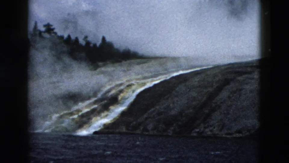 1958 Yellowstone home-movie still showing steam over a dark rocky slope, water or runoff below, and distant evergreen trees under cloudy sky.