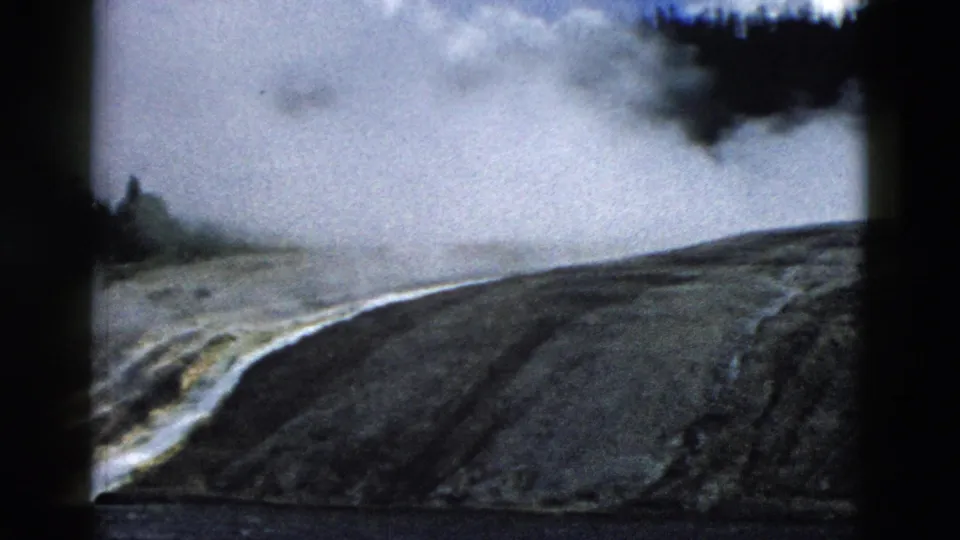 1958 Yellowstone frame showing a dark mineral slope, rising steam, water or runoff at the bottom, and distant trees softened by mist.