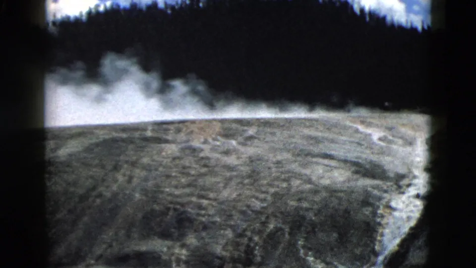Final 1958 Yellowstone frame with steam spreading near the forest edge over a mineral-streaked geothermal hillside.