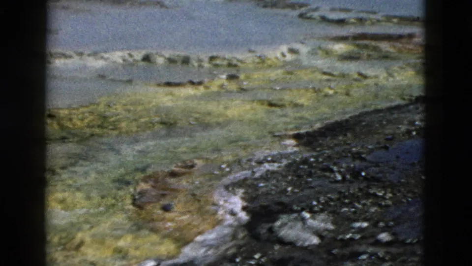 1958 Yellowstone 8mm frame showing a wider geothermal pool surface, green mineral mats, pale deposits, and a dark rocky bank in the foreground.