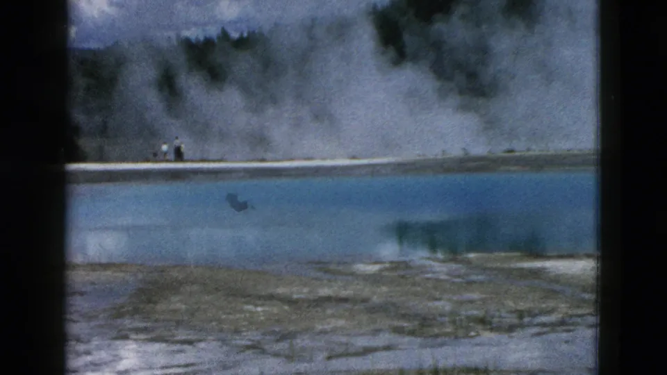 1958 Yellowstone home-movie frame with distant visitors on a pale shore beside heavy geothermal steam.