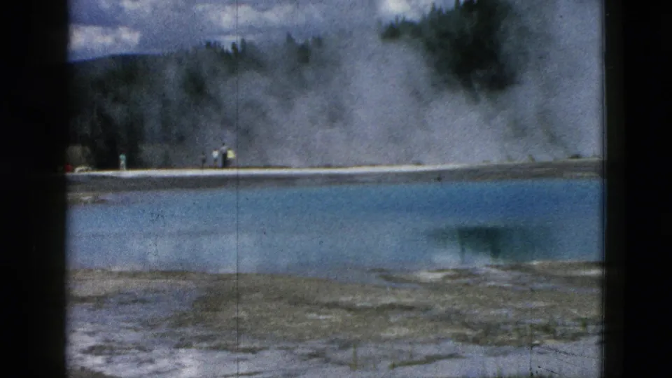1958 Yellowstone frame showing distant visitors along a pale geothermal shore, with blue water and steam behind them.