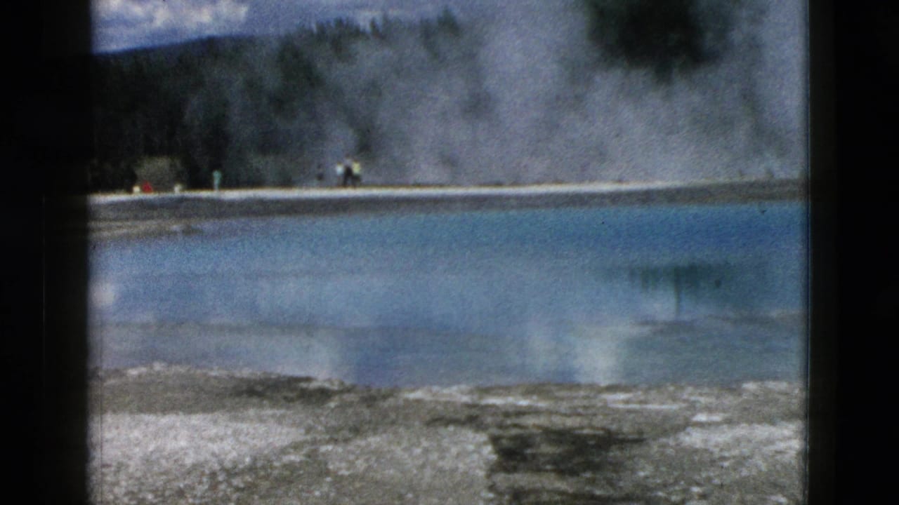 Steaming blue hot spring bordered by pale mineral ground and distant visitors near the far shore.
