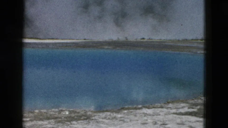 1958 Yellowstone frame showing a steaming blue hot spring, white and gray mineral edges, distant treeline, and soft 8mm film texture.
