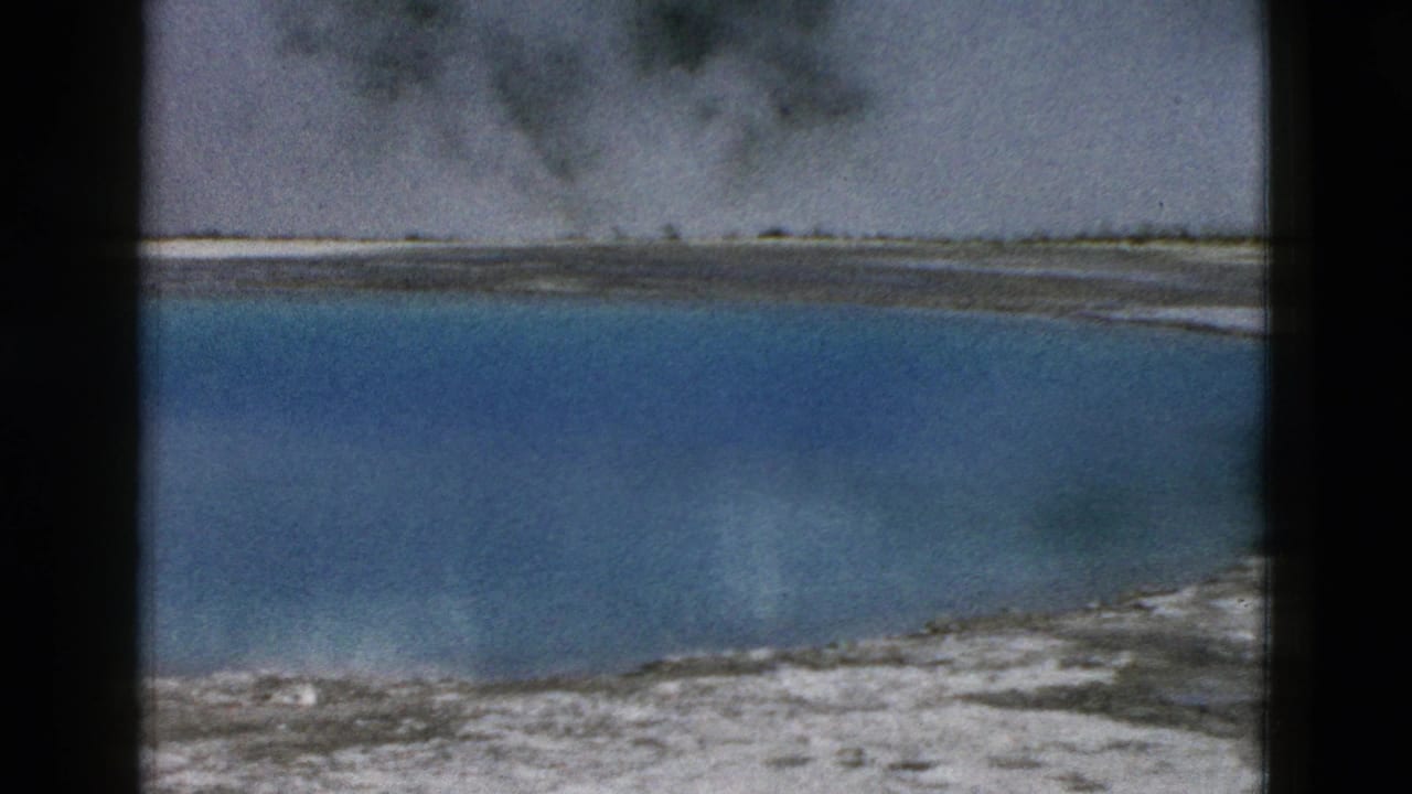 Blue geothermal pool with pale mineral edge, steam, trees, and cloudy reflection.