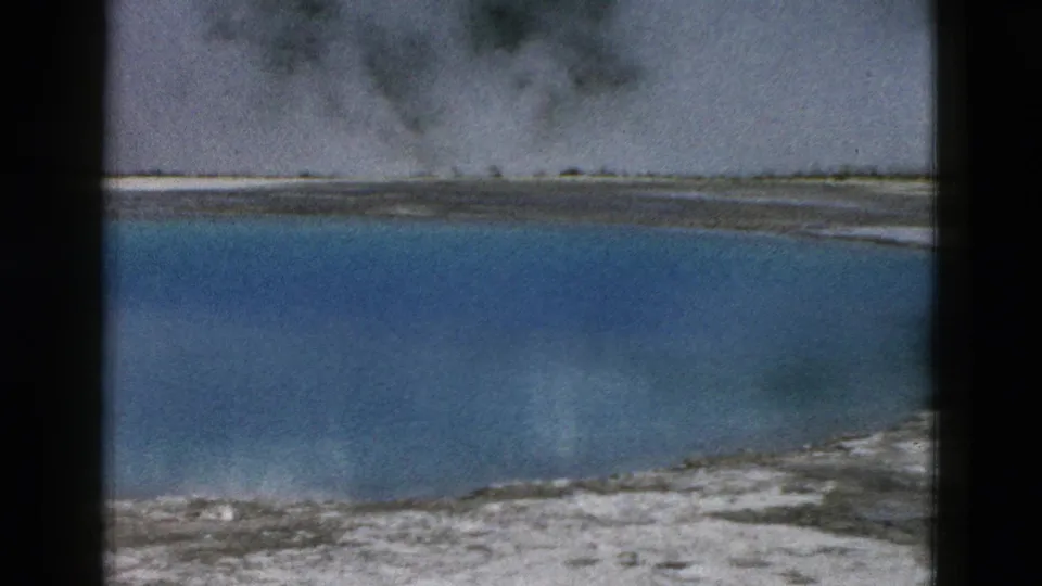 1958 Yellowstone 8mm frame showing a blue geothermal pool, pale mineral shoreline, steam, and cloud reflections.