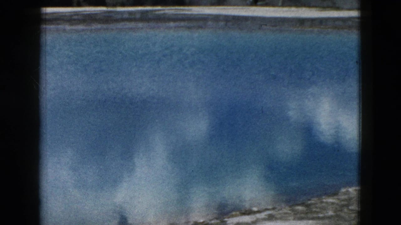 Yellowstone geothermal pool surface in 1958 home-movie footage, with blue water, reflected clouds, and a pale rocky edge.
