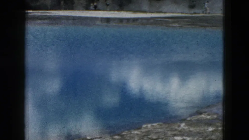 8mm Yellowstone view of a blue geothermal pool with cloud reflections and a light mineral shoreline in the foreground.