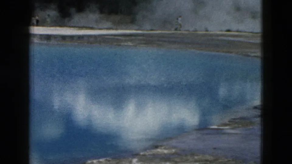 1958 Yellowstone frame with a distant visitor walking along the mineral edge of a blue geothermal pool with steam behind.