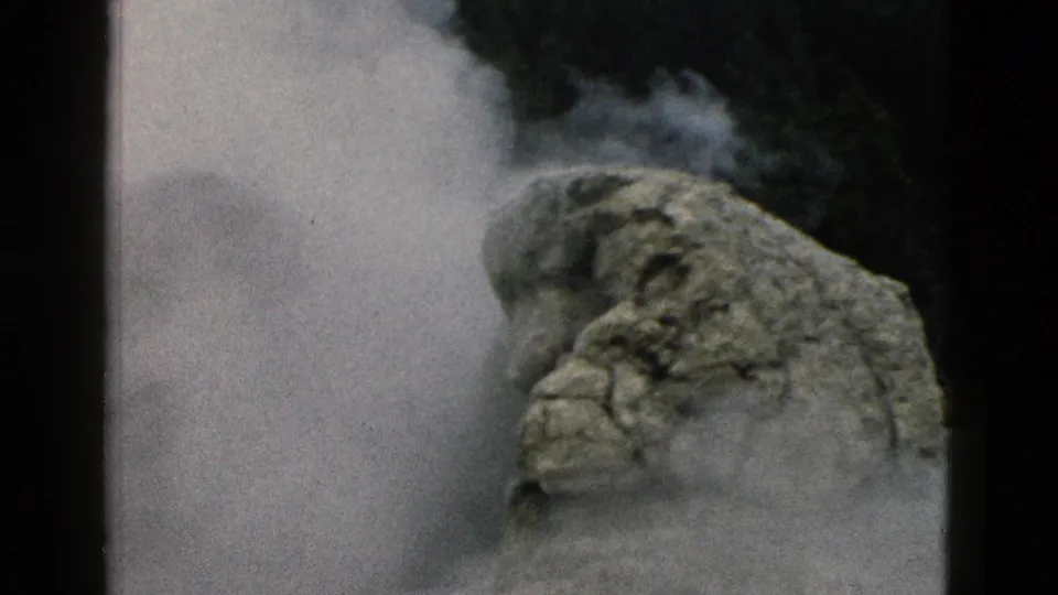 A white steam plume moves behind a large rough rock formation in Yellowstone, framed by dark 8mm gate edges and soft archival grain.