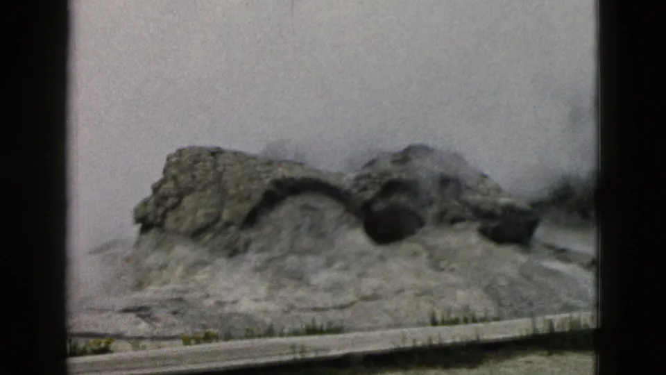 1958 Yellowstone home-movie frame showing steam drifting from the Old Faithful geyser mound with a low barrier in front.