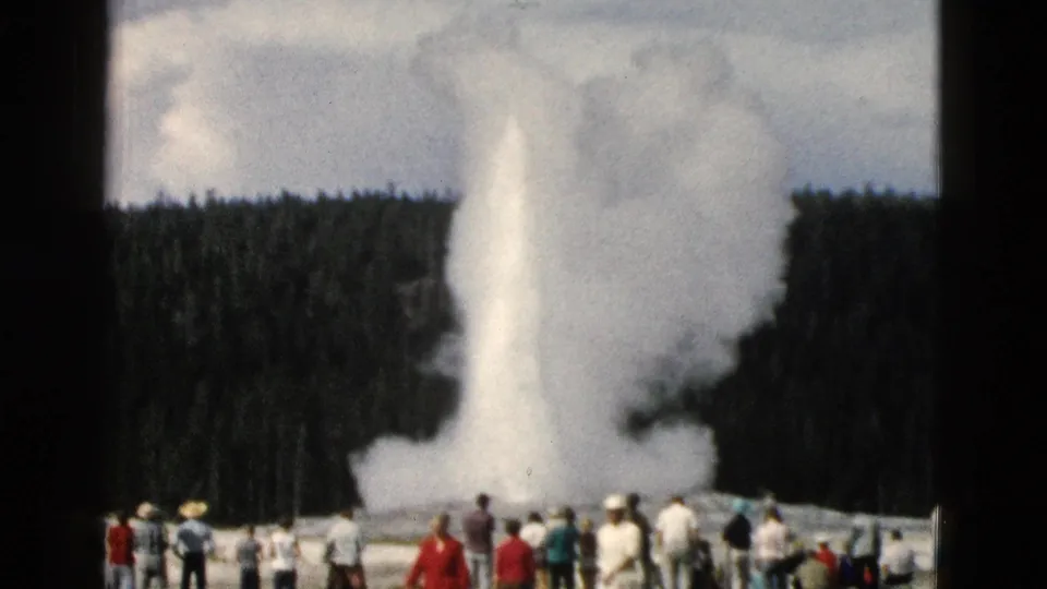 1958 Yellowstone frame showing Old Faithful erupting while a distant crowd of tourists watches from the viewing area.