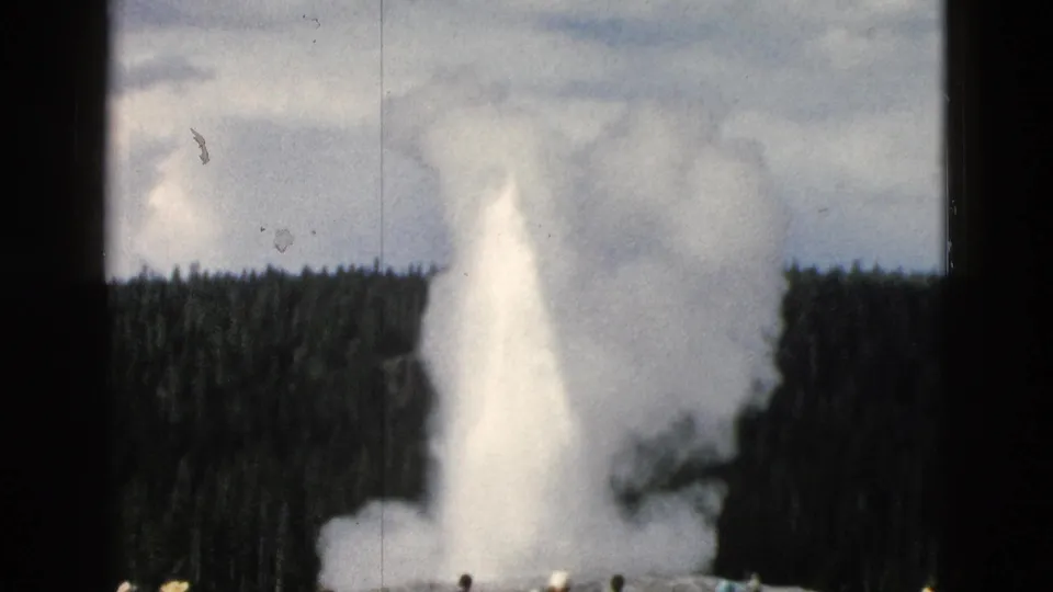 1958 Yellowstone home-movie frame showing Old Faithful erupting with steam, pine forest, cloudy sky, and tourists near the base.