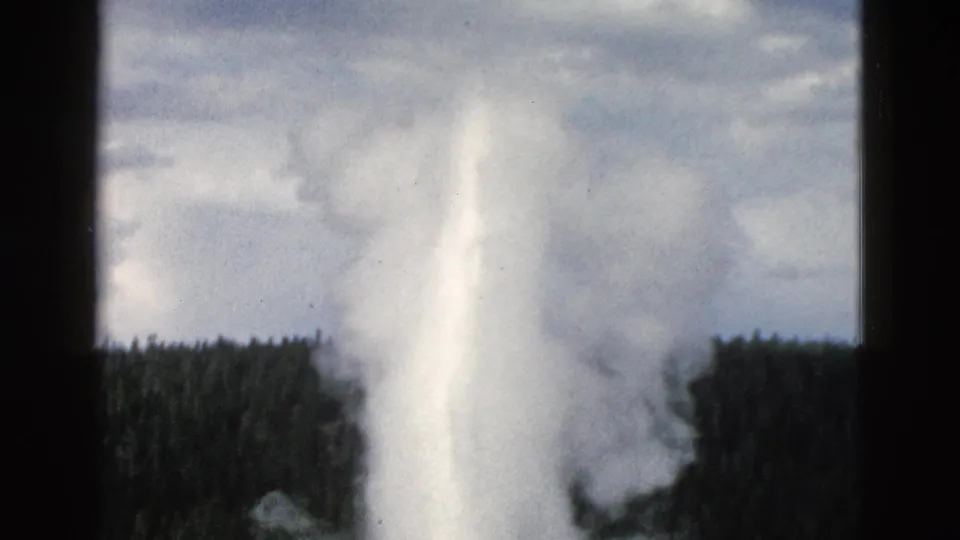 1958 Yellowstone frame with Old Faithful’s plume filling the view, steam drifting across dark trees and a cloudy sky.