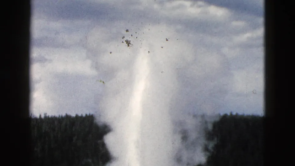 Yellowstone 8mm frame showing Old Faithful’s white plume and steam cloud with pine forest, cloudy sky, and visible film dust.