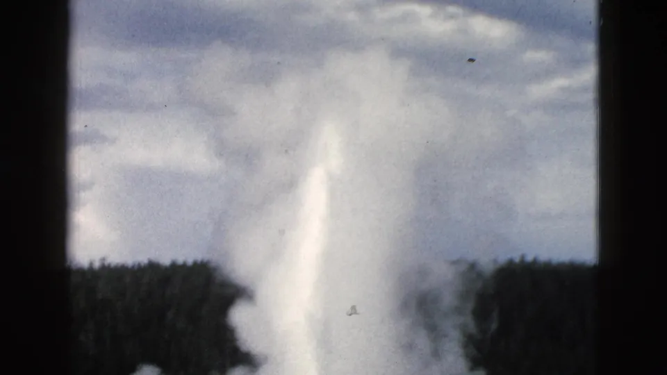 1958 Yellowstone 8mm frame showing a geyser eruption rising above forest silhouettes beneath overcast clouds.