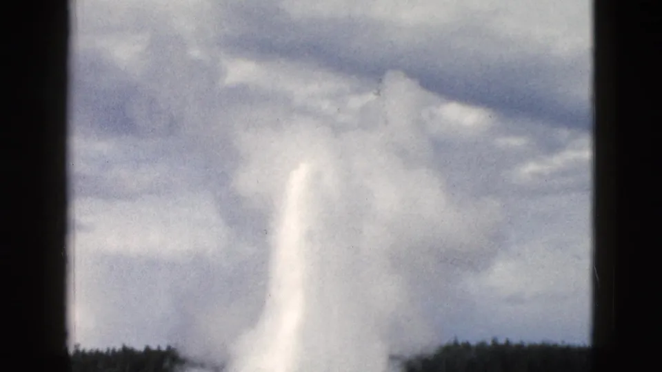 1958 Yellowstone 8mm frame showing geyser spray and steam rising into a pale cloudy sky above forest silhouettes.