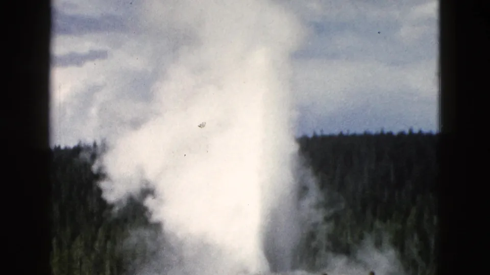 A bright Old Faithful eruption column fills the lower frame, with a dark forested horizon and cloudy sky in Yellowstone.