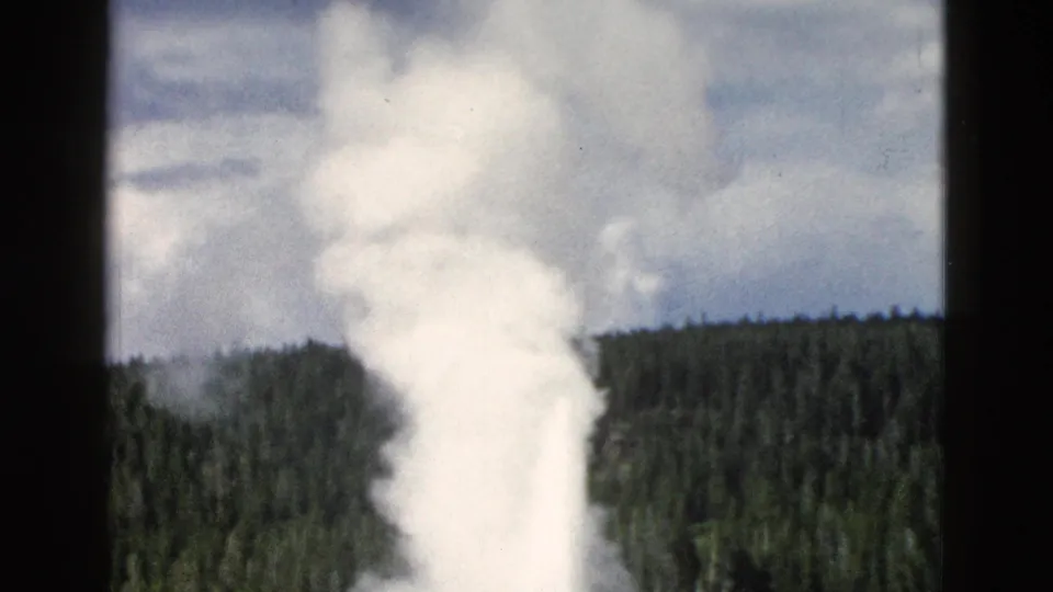 Old Faithful erupts in Yellowstone as small distant tourists watch near the lower edge of the 8mm frame.