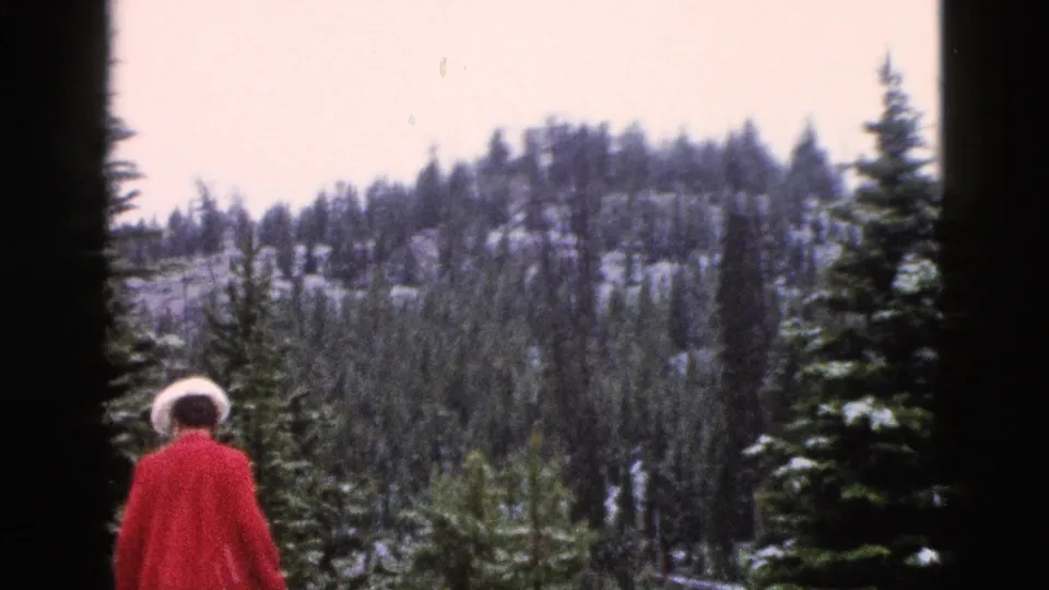 1958 Yellowstone frame with a woman in a red coat and white hat walking away among snowy evergreens and distant wooded slopes.