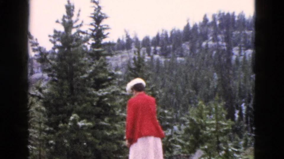 1958 Yellowstone 8mm frame showing the woman in a red coat moving past a tall evergreen in a snow-dusted forest.