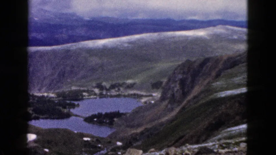 1958 8mm travel footage frames a high mountain valley with blue lakes, rocky slopes, scattered snow patches, and distant ridges.
