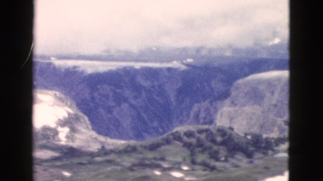 1958 Yellowstone home-movie frame showing a deep valley, rugged cliff faces, lingering snow, and hazy sky from a distant viewpoint.