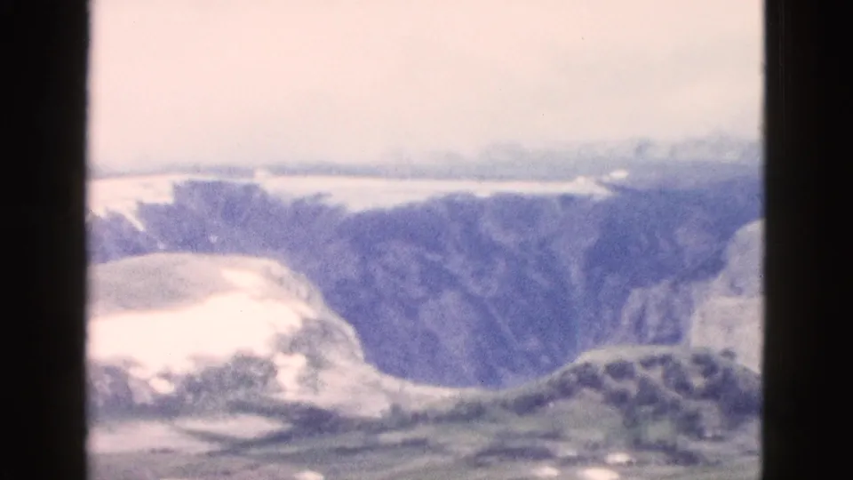 1958 Yellowstone frame showing a hazy mountain valley with snow patches, steep cliff texture, and foreground terrain in soft 8mm focus.