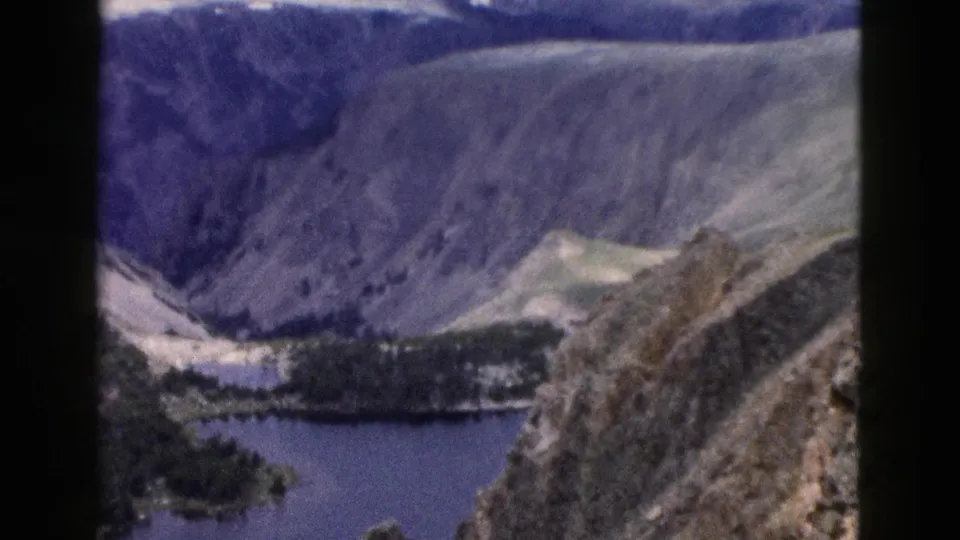 1958 Yellowstone-area scenic view of a blue lake set below rugged mountain slopes, with evergreen stands and pale meadow areas visible in the valley.
