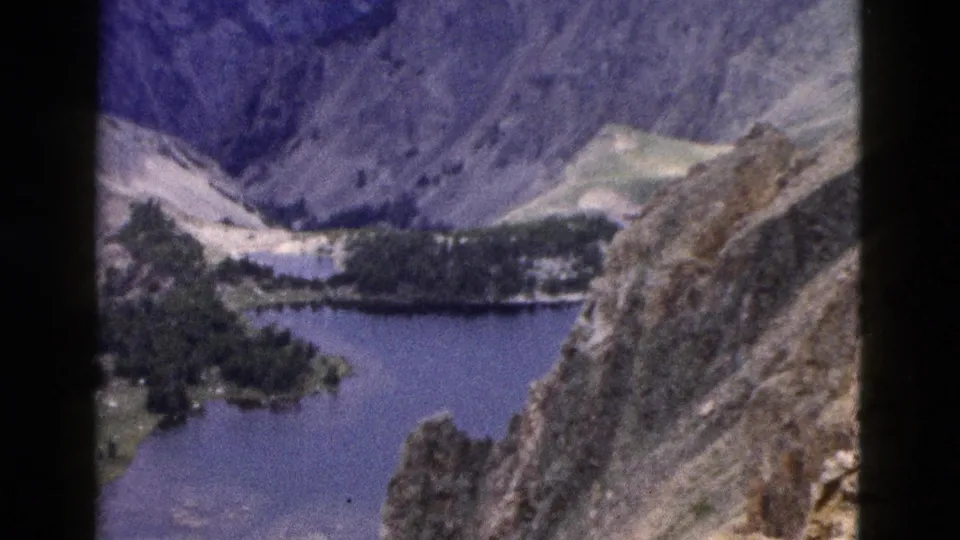 1958 Yellowstone-area 8mm frame showing a broad mountain valley with lake water, evergreen clusters, rocky foreground terrain, and muted ridges beyond.
