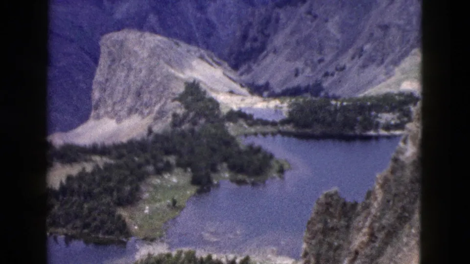1958 Yellowstone-area mountain panorama with a rocky peak, blue lake basin, evergreen shoreline, meadow patches, and steep ridges in soft 8mm color.