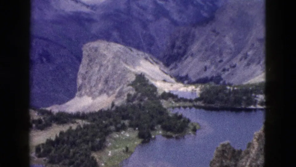 1958 Yellowstone home-movie frame showing a rocky mountain, alpine lake, and evergreen slopes from a distant overlook.