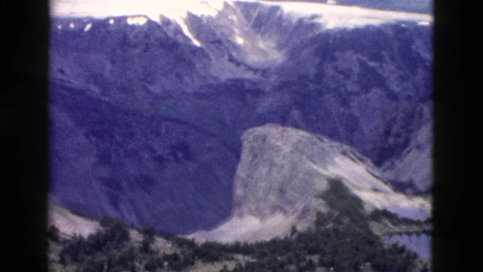 1958 Yellowstone landscape frame with a snow-streaked ridge and pale rocky outcrop above dark forested terrain.