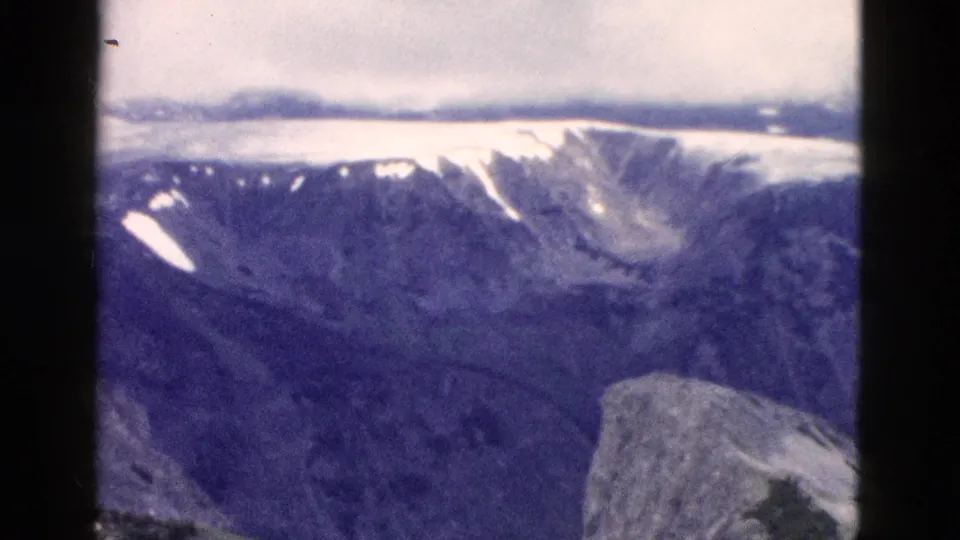1958 Yellowstone home-movie view across a broad mountain basin with lingering snow patches and a rocky foreground edge.