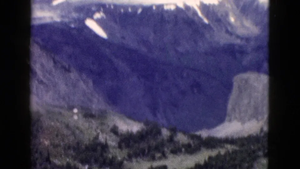 1958 Yellowstone frame showing a shadowed mountain slope, snow patches, evergreen forest, and part of a pale rocky outcrop.