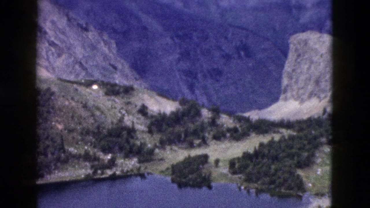 1958 Yellowstone home-movie frame showing a blue alpine lake, forested shoreline, and pale rock faces beneath steep mountains.