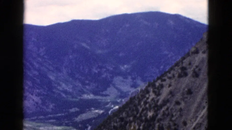 1958 Yellowstone home-movie view looking across a deep mountain valley, with a steep rocky slope in the foreground and blue hills beyond.
