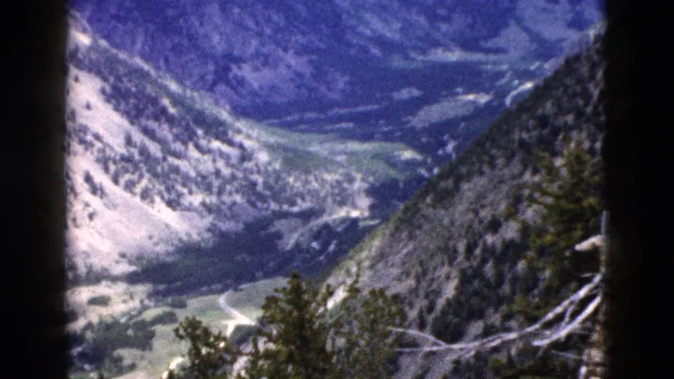 1958 Yellowstone 8mm frame with a rocky slope, evergreen-covered ridge, and winding road across the valley below.
