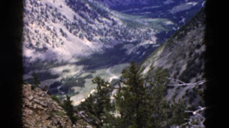 1958 8mm Yellowstone frame looking past rocky foreground and trees toward a winding road through a mountain valley.