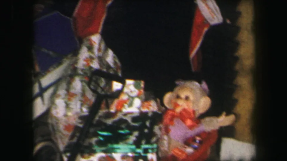 1971 Lynbrook home-movie frame of Christmas gifts, a doll stroller, and a plush monkey toy under a decorated tree.