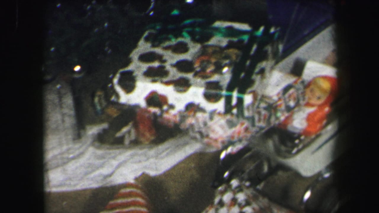 1971 Lynbrook home-movie frame showing wrapped holiday gifts, a small doll-like figure, and patterned fabric in a dim interior.