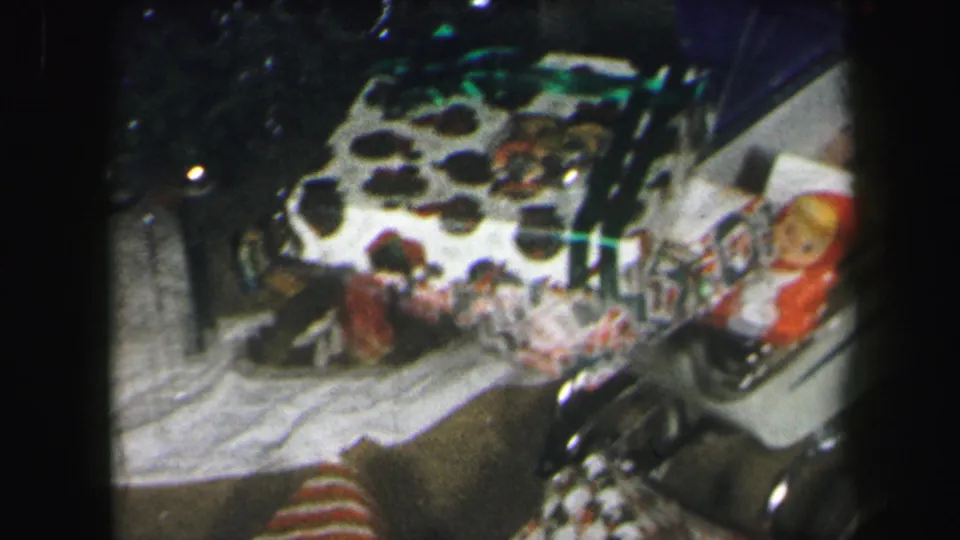 1971 Lynbrook home-movie frame showing wrapped holiday gifts, a small doll-like figure, and patterned fabric in a dim interior.